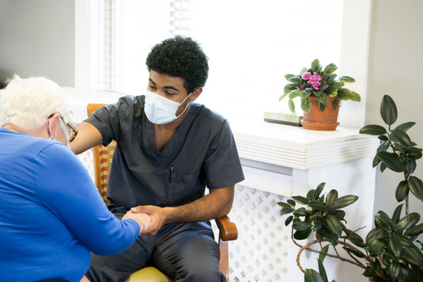 Portrait of a smling African-American male nursing employee wearing gray scrubs and face mask interacts with a senior aged patient during the Covid-19 pandemic, Midwest, USA