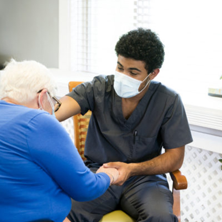 Portrait of a smling African-American male nursing employee wearing gray scrubs and face mask interacts with a senior aged patient during the Covid-19 pandemic, Midwest, USA